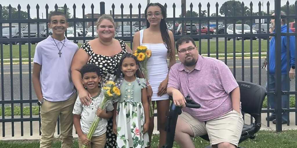 Matt Curcio and his family pose outdoors by a fence, with the children holding sunflowers and Matt seated on a mobility scooter.