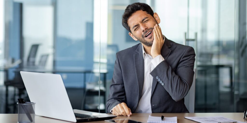 Arab businessman suffering from toothache, sitting in the office at the table in front of the laptop, holding his jaw, grimacing in pain, health concept.