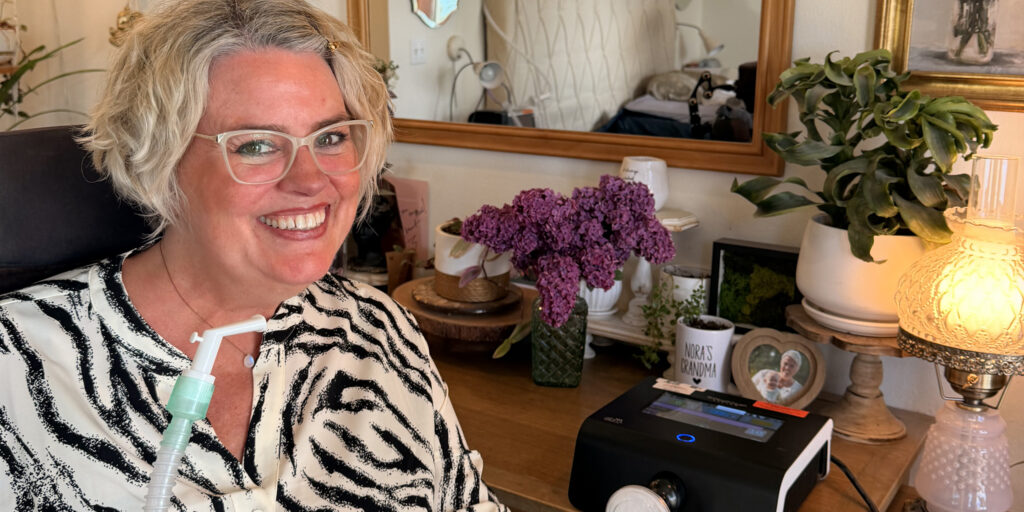 Woman smiling beside a home ventilator in a cozy room with flowers and houseplants.