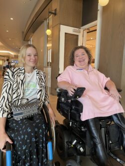Two women in power wheelchairs pose in a hotel hallway