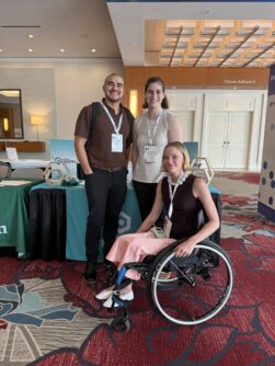 A young woman in a wheelchair poses next two a man and woman standing by a table at a conference