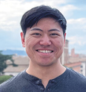 Headshot of MDA’s Research Portfolio Director Brian Lin smiling at the camera outdoors against a background of adobe-colored buildings and a blue sky.