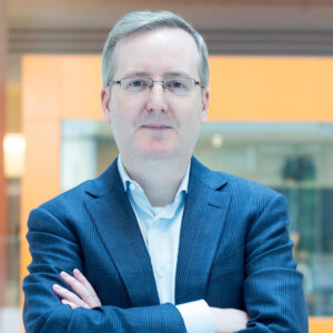 Headshot of researcher Bryan Traynor wearing glasses and a blue blazer posing with arms crossed in a professional indoor setting.