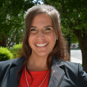 Headshot of Danica Gonzalves in a black jacket and red shirt, smiling at the camera with green trees in the background.