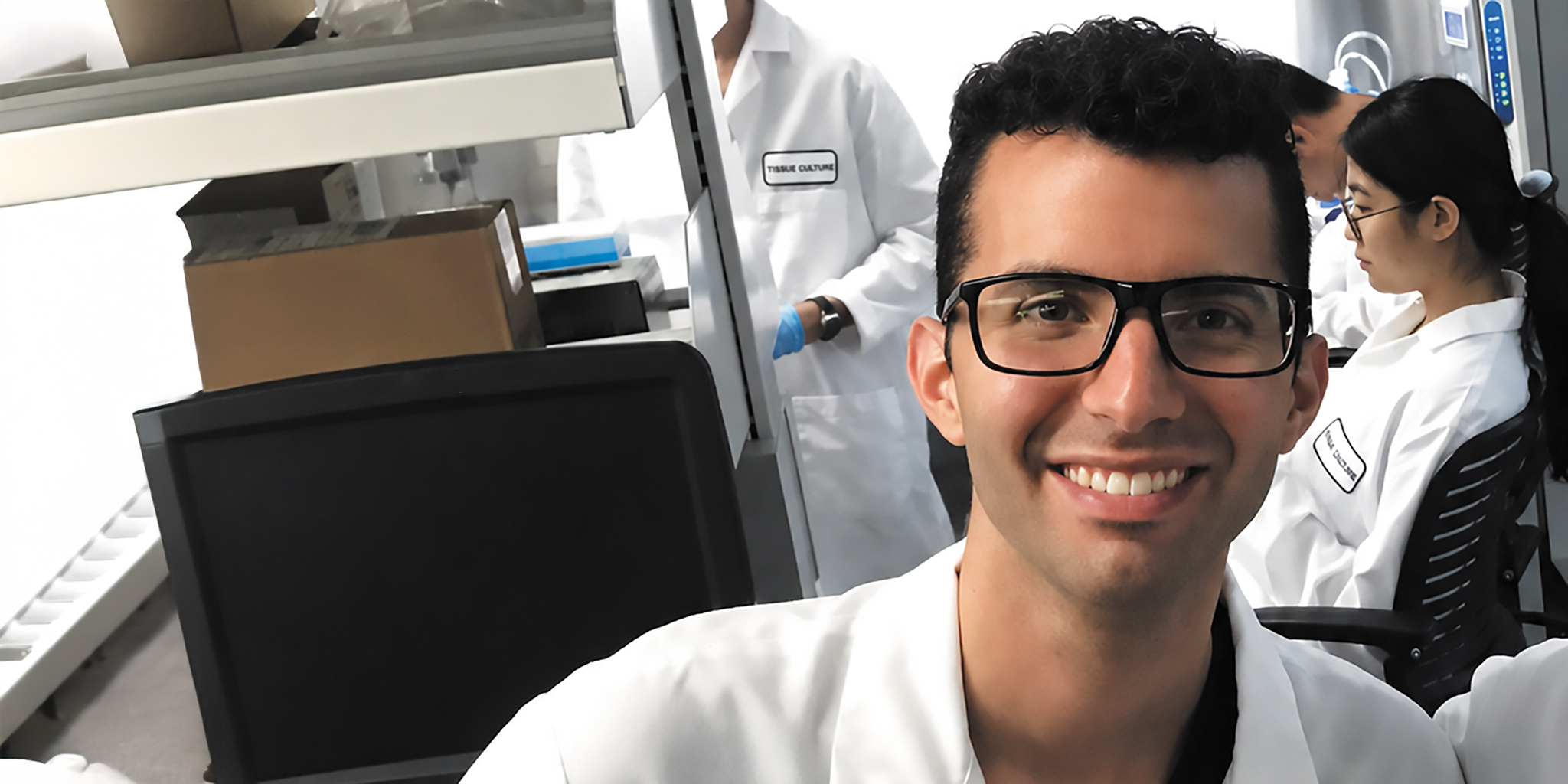 Bioengineer Ioannis (John) Eugenis wearing glasses and a white lab coat in a laboratory with colleagues.