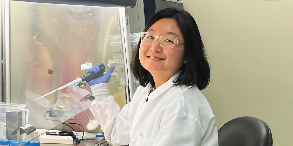 Researcher Asuka Eguchi in a white lab coat smiles while holding a pipette at a laboratory workstation.
