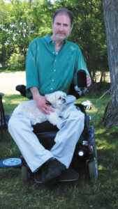 Attorney Ed Barocas sits in a power wheelchair holding a small white dog in his lap outdoors next to a tree.