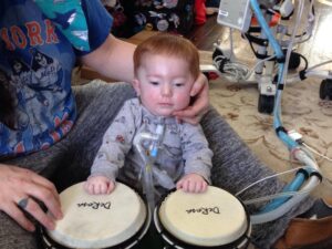 Stevie, at 1 year old, seated in front of a pair of bongos supported by a caregiver with a tracheostomy and medical equipment in the background.