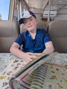 Stevie, at 8 years old, wearing a baseball cap sits at a table in the cabin of a ferry boat with an open sticker book.