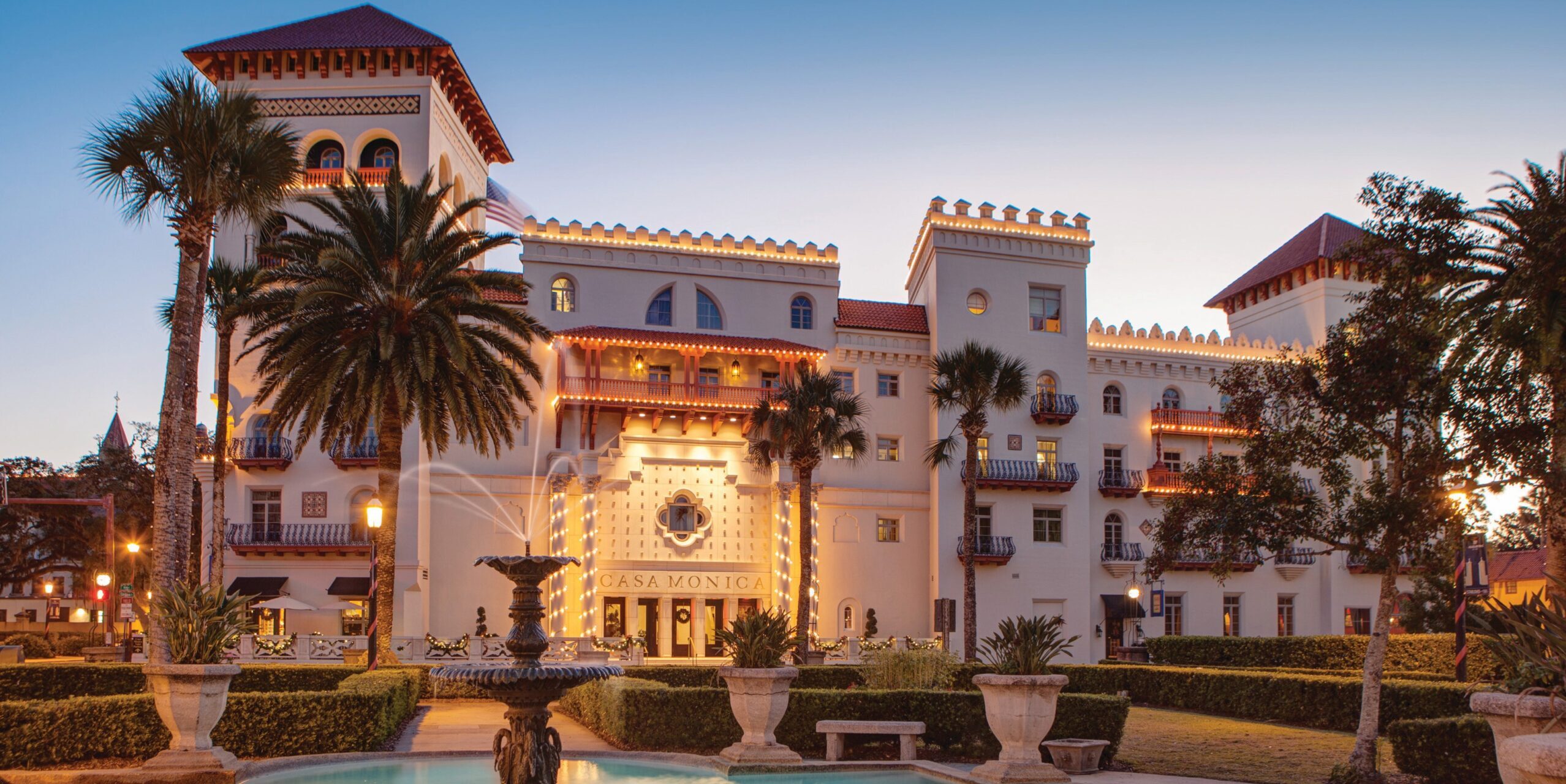 Historic Moorish Revival style hotel with palm trees and a fountain illuminated at dusk.
