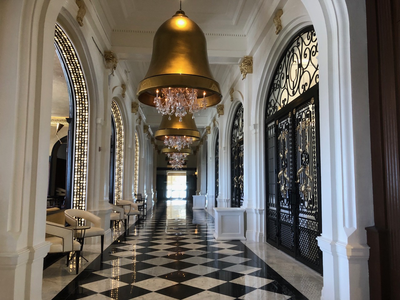 Ornate hotel corridor with black and white marble flooring and crystal chandeliers encased in gold bell-shaped covers.
