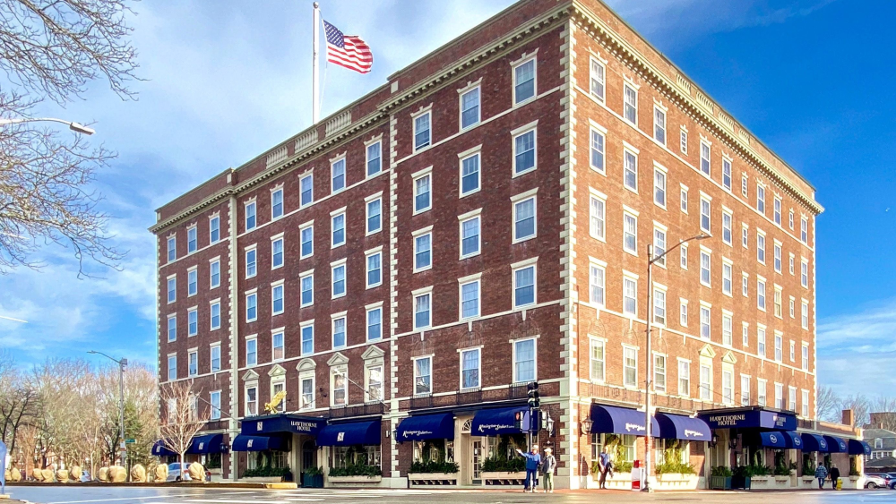 Red brick and stone Federal style hotel building with a large American flag on top and blue awnings on the ground floor.
