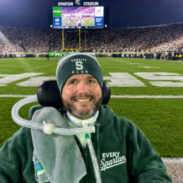 A man in a wheelchair with a ventilator smiles at a college football game