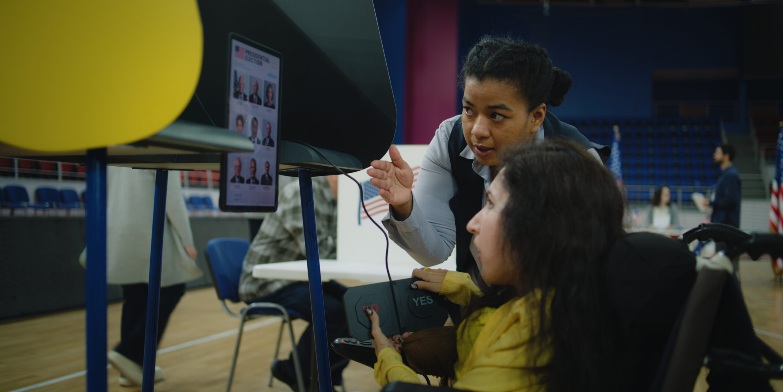 Polling officer consults woman with physical disability. Female US voter with SMA uses remote buttons and votes on tablet computer in voting booth at polling station. Election Day in United States.