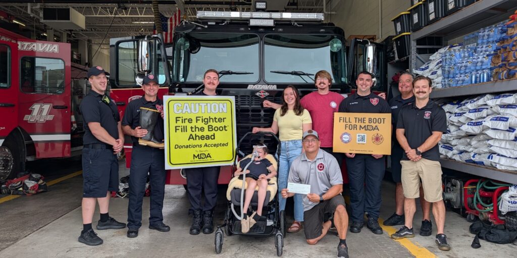 Fire fighters pose with an MDA Ambassador in front of a fire truck while holding Fill the Boot signs
