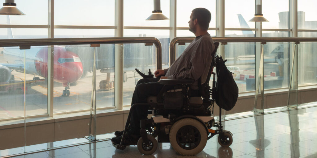 Person with disability using wheelchair observes planes through large windows at an airport terminal.